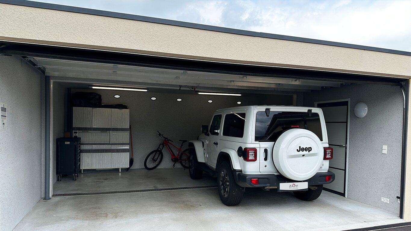 Betonfertiggarage mit geöffnetem Sektionaltor, SUV und Stauraum Fertiggarage mit geöffnetem Sektionaltor, weißer SUV geparkt rechts, links Schrank und Regalsystem sowie Fahrrad im Hintergrund, integrierte Beleuchtung an der Decke, von ZAPF