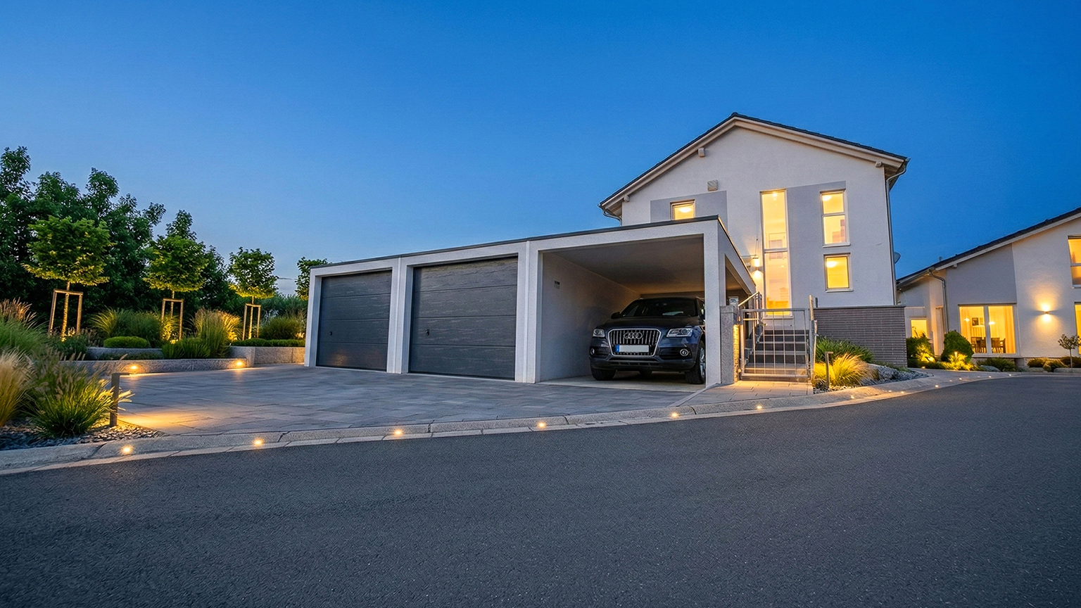 Doppelgarage aus Beton mit Sektionaltoren und integriertem Carport am Einfamilienhaus Moderne Betonfertiggarage mit zwei grauen Sektionaltoren und seitlichem Carport, davor gepflasterte Einfahrt mit Beleuchtung, ein Fahrzeug unter dem Carport, angrenzend ein beleuchtetes Einfamilienhaus in Hanglage von ZAPF