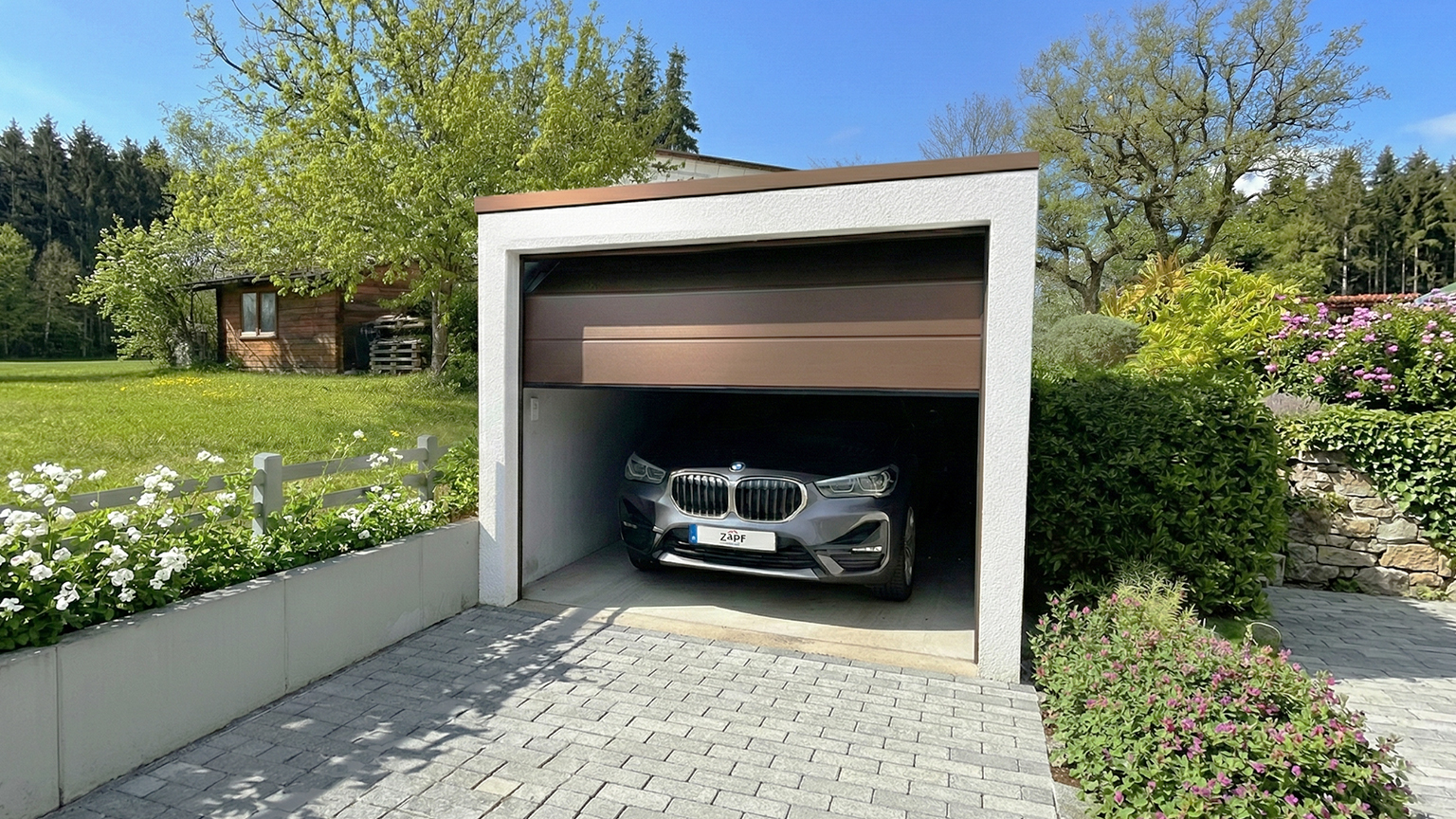 Kompakte Betonfertiggarage mit halb geöffnetem Sektionaltor im Garten Freistehende Betonfertiggarage mit teilweise geöffnetem braunem Sektionaltor und geparktem Fahrzeug im Inneren, davor gepflasterte Einfahrt, umgeben von Blumenbeeten, Hecken und Bäumen in gepflegter Gartenanlage von ZAPF
