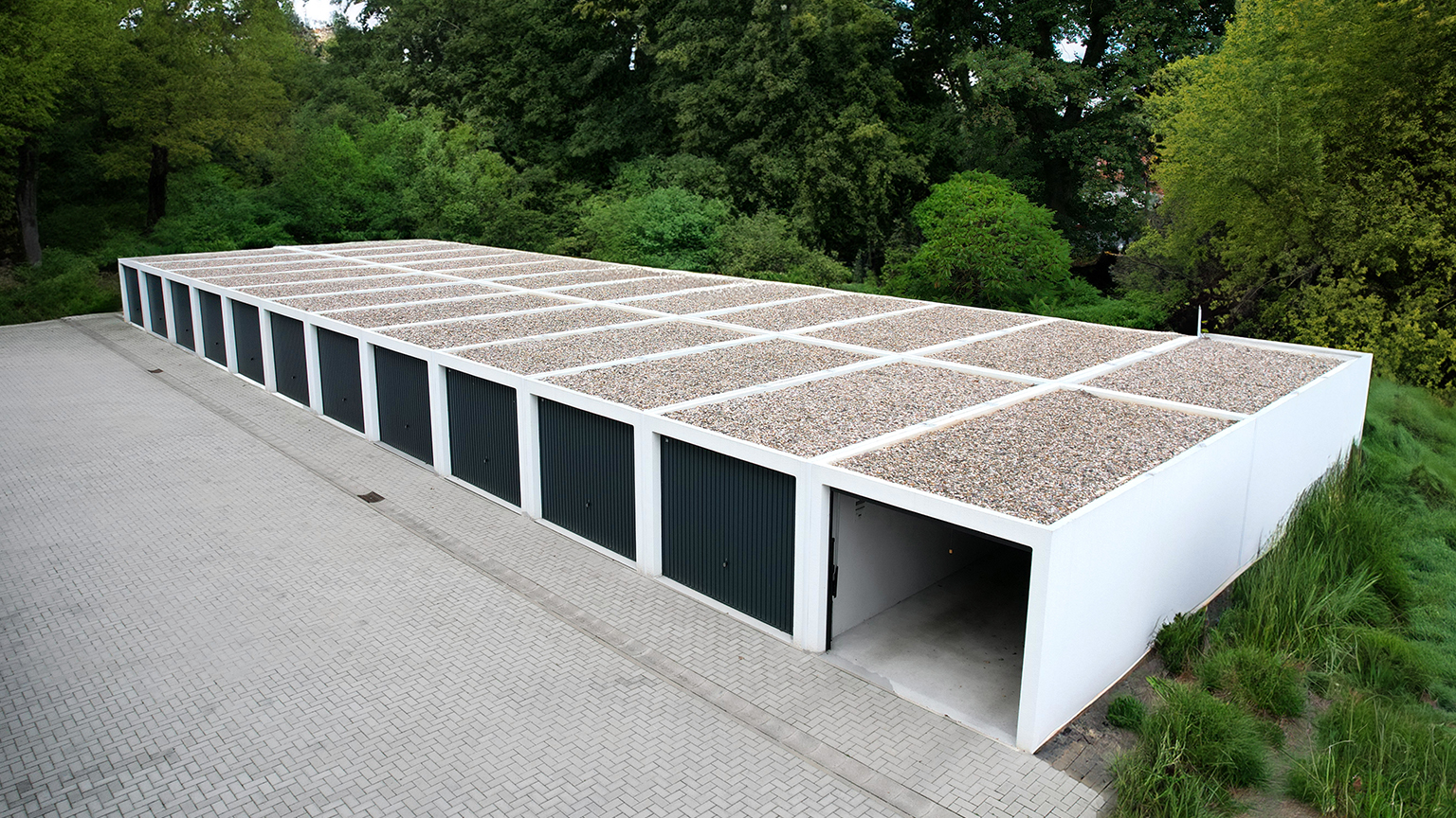 Reihengaragen aus Betonfertiggaragen mit Flachdach und Kiesauflage in grüner Umgebung Lange Reihe weißer Betonfertiggaragen mit Flachdächern und Kiesauflage sowie dunklen Garagentoren entlang einer gepflasterten Zufahrt, umgeben von Bäumen und Grünflächen von ZAPF