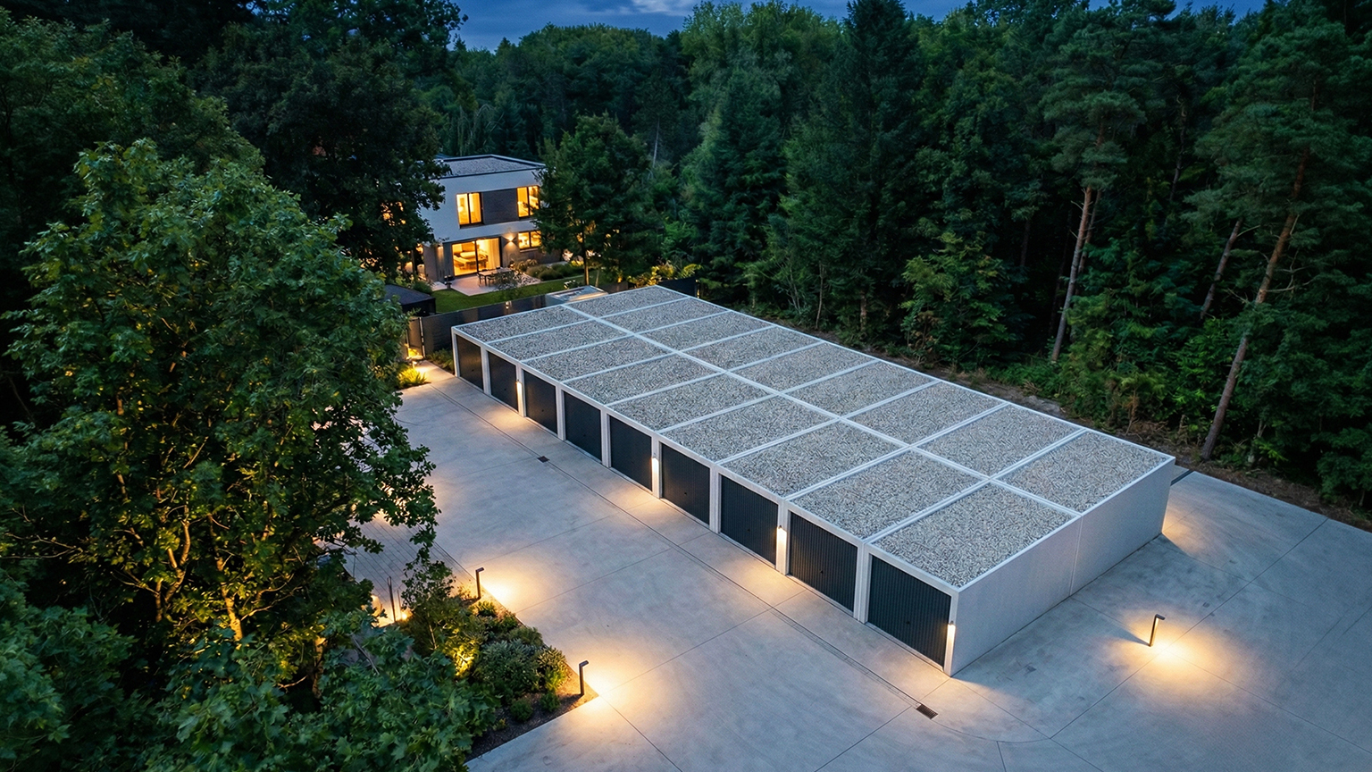 Große Betonfertiggaragenanlage mit Dachbegrünung neben modernem Wohnhaus im Wald Mehrere aneinandergereihte Betonfertiggaragen mit dunklen Garagentoren und extensiv begrüntem Flachdach auf einem beleuchteten Hof neben einem modernen Wohnhaus, umgeben von dichtem Baumbestand in der Dämmerung von ZAPF Garagen
