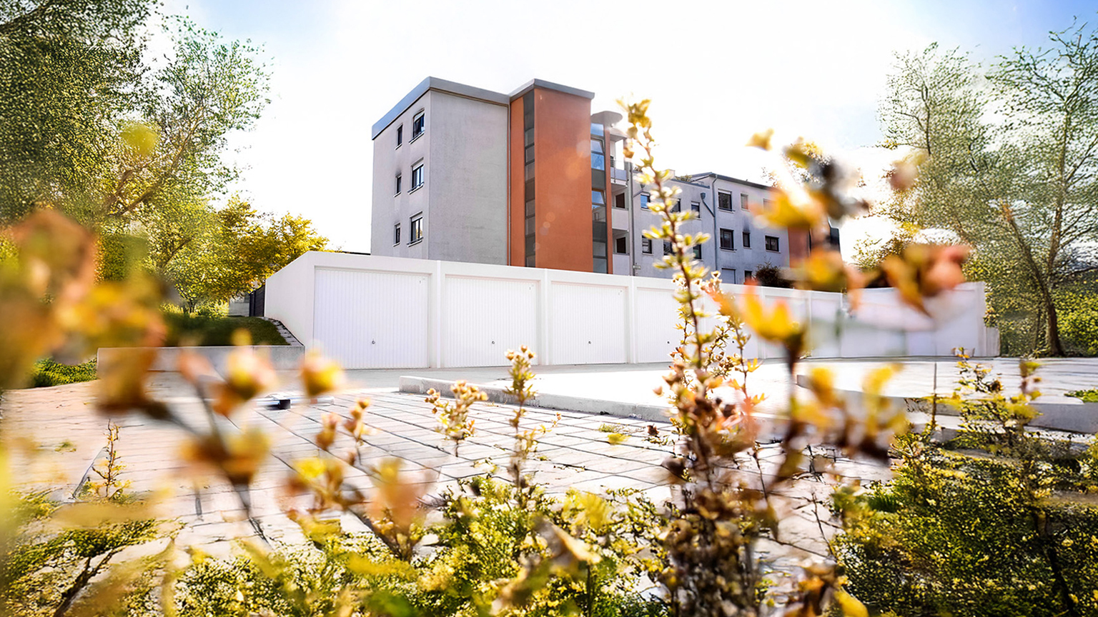 Weiße Betonfertiggaragen mit Kipptoren vor Mehrfamilienhaus in grüner Umgebung Reihe von weißen Betonfertiggaragen mit hellen Kipptoren auf gepflasterter Fläche vor einem modernen Mehrfamilienhaus, umgeben von Bäumen und bepflanzten Bereichen im Sonnenlicht von ZAPF Garagen
