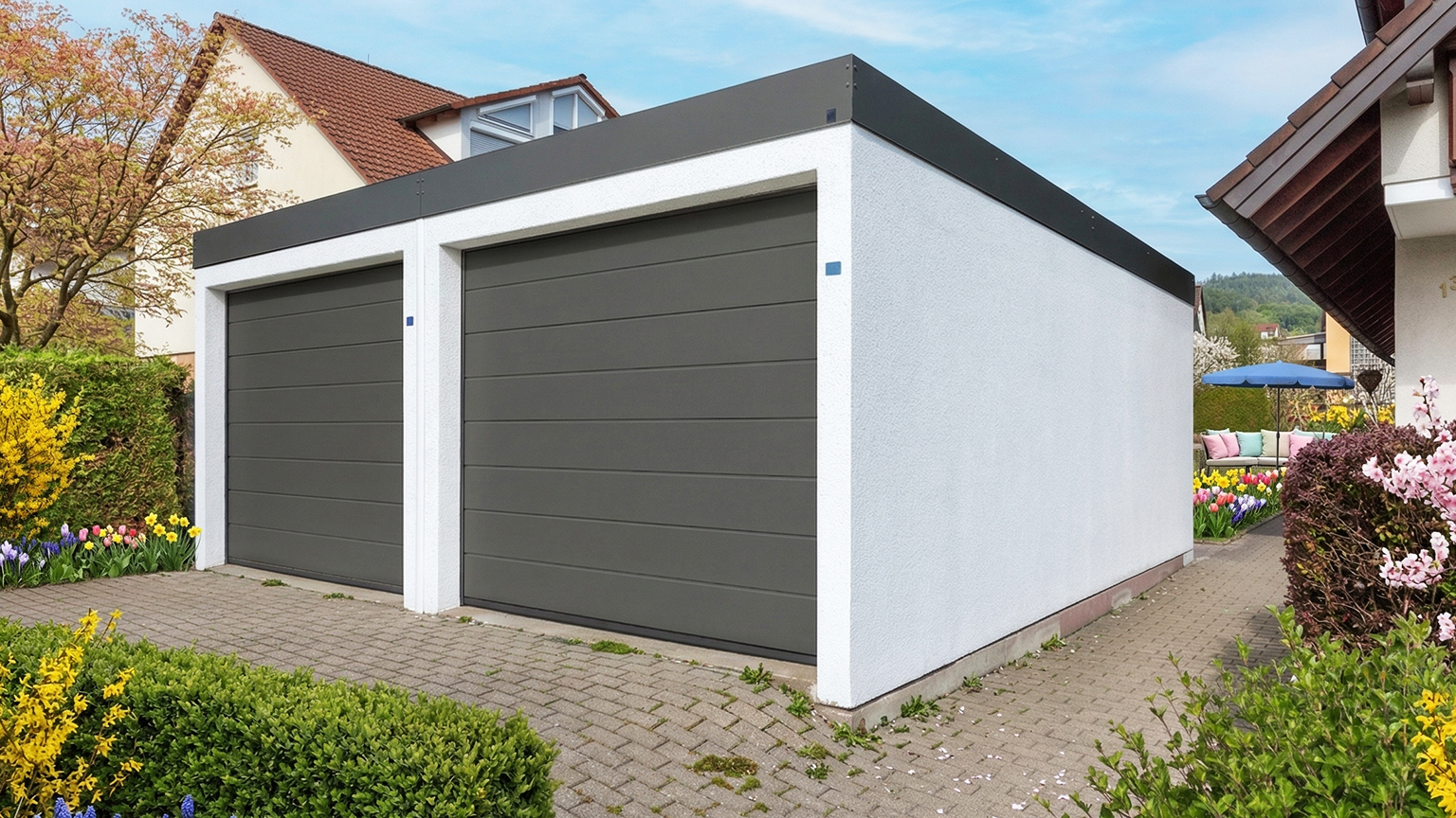 Moderne Doppel Fertiggarage mit grauen Sektionaltoren in Wohngebiet Freistehende Betonfertiggarage mit zwei breiten grauen Sektionaltoren, weiß verputzter Fassade und Flachdach in gepflegter Gartenumgebung mit Pflasterfläche und Blumenbeeten von ZAPF