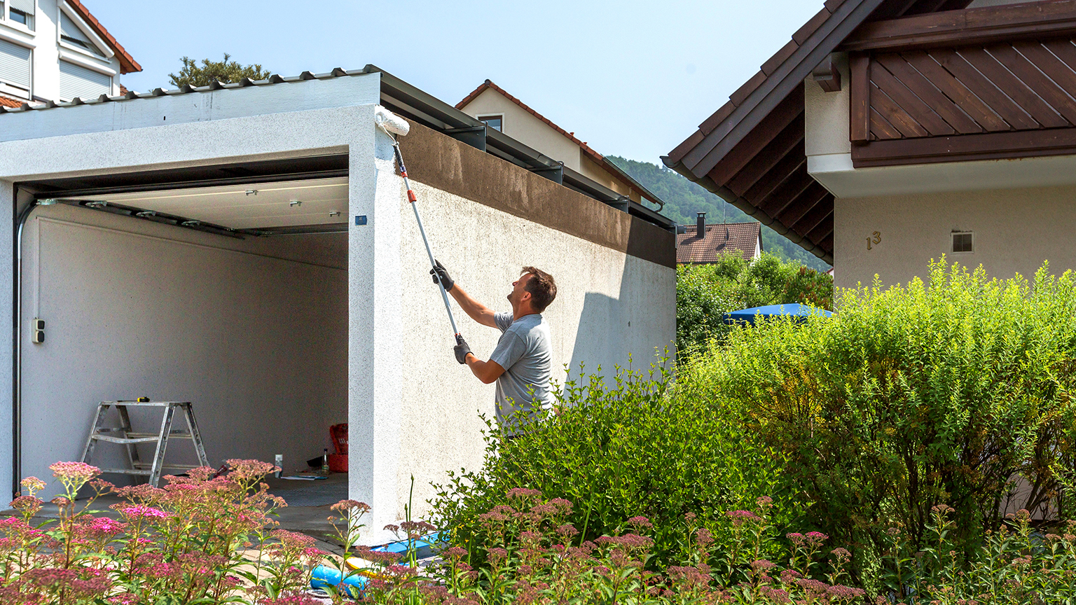 Pflege und Anstrich einer Betonfertiggarage mit Farbrolle Mann streicht die Fassade einer Betonfertiggarage mit einer Farbrolle an der Außenwand, offene Garage mit Leiter im Inneren, umgeben von Gartenpflanzen in Wohngebiet von ZAPF