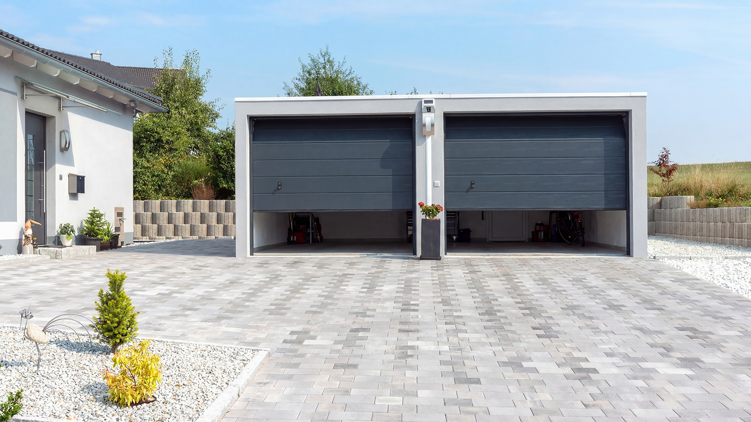 Doppelgarage mit geöffneten Sektionaltoren und großzügiger Pflasterfläche Moderne Betonfertiggarage mit zwei teilweise geöffneten Sektionaltoren, großer gepflasterter Einfahrt und aufgeräumtem Vorplatz neben Einfamilienhaus und Gartenanlage von ZAPF