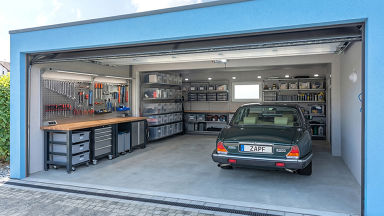 Offene Fertiggarage mit Sektionaltor, aufgeräumter Werkstattwand, Werkbank und Regalsystemen sowie einem abgestellten Pkw im Innenraum von ZAPF