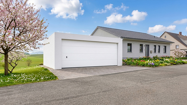 Weiße Betonfertiggarage mit breitem Sektionaltor auf gepflasterter Zufahrt vor einem modernen Einfamilienhaus, umgeben von gepflegtem Garten mit Blumenbeeten und blühendem Baum von ZAPF