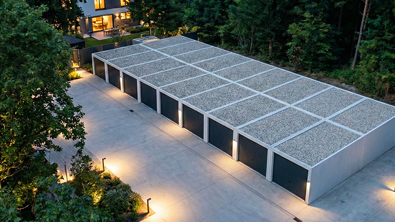 Mehrere aneinandergereihte Betonfertiggaragen mit dunklen Garagentoren und extensiv begrüntem Flachdach auf einem beleuchteten Hof neben einem modernen Wohnhaus, umgeben von dichtem Baumbestand in der Dämmerung von ZAPF Garagen