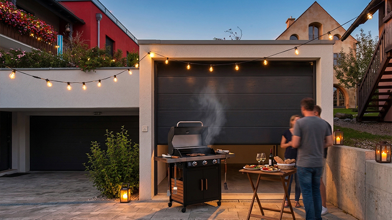 Moderne Betonfertiggarage mit geöffnetem Sektionaltor bei Abenddämmerung, integrierter Grill im Innenraum, beleuchtete Lichterkette, gedeckter Beistelltisch und Personen im Gespräch vor der Garage, von ZAPF