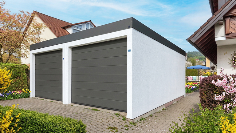 Freistehende Betonfertiggarage mit zwei breiten grauen Sektionaltoren, weiß verputzter Fassade und Flachdach in gepflegter Gartenumgebung mit Pflasterfläche und Blumenbeeten von ZAPF