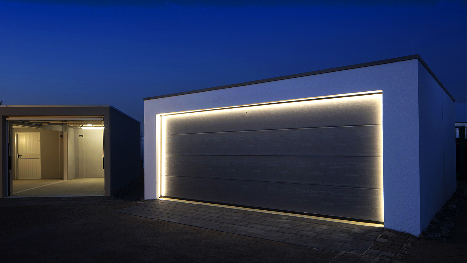 Precast concrete garage with illuminated sectional door at dusk Modern precast concrete garage with a large dark sectional door, surrounding LED lighting and an open single garage beside it by ZAPF
