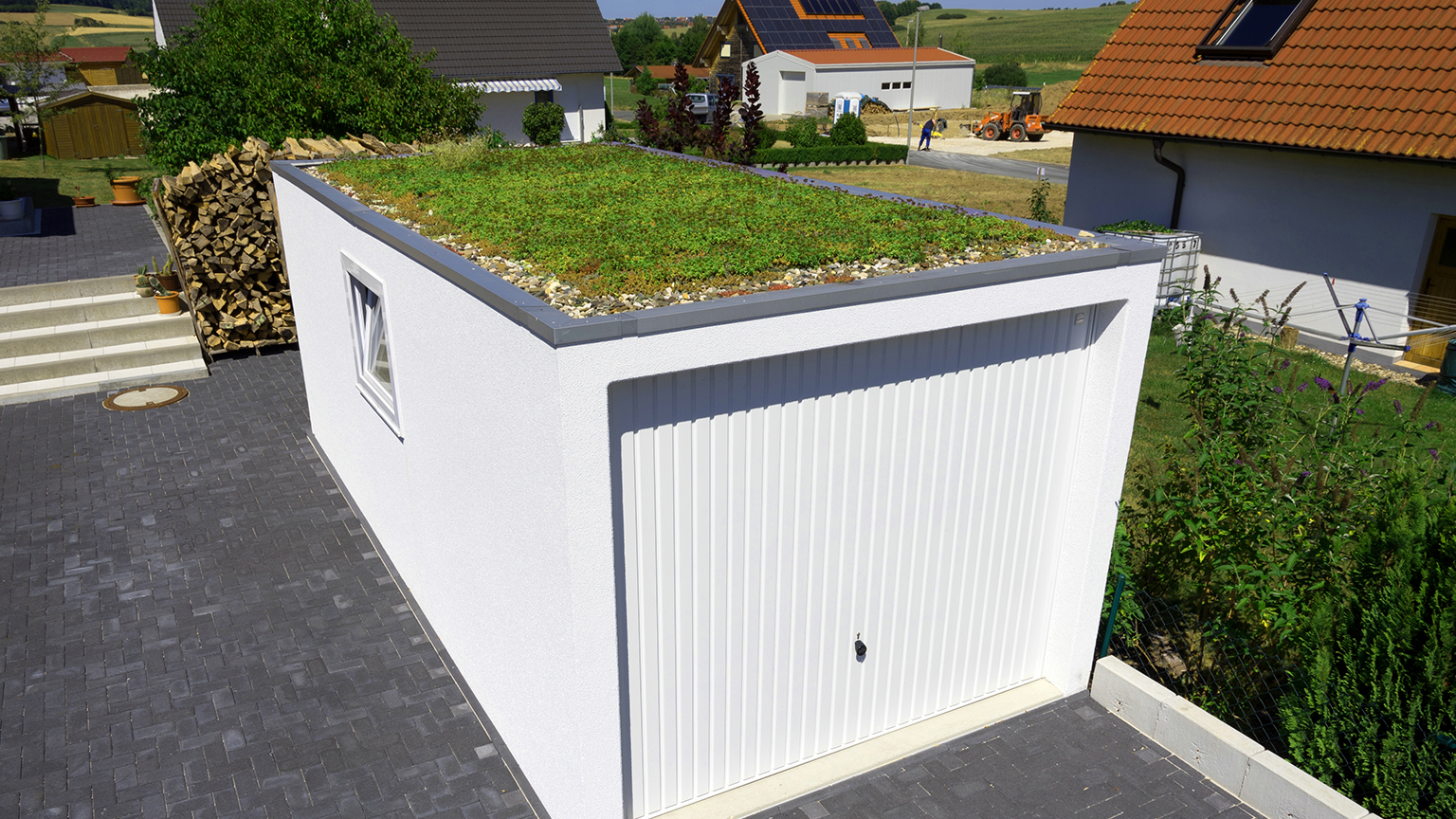 Garage préfabriqué blanc avec toiture végétalisée et porte basculante Garage préfabriqué blanc avec toit plat végétalisé, bordure de gravier, fenêtre latérale et porte de garage blanche sur une allée pavée de ZAPF