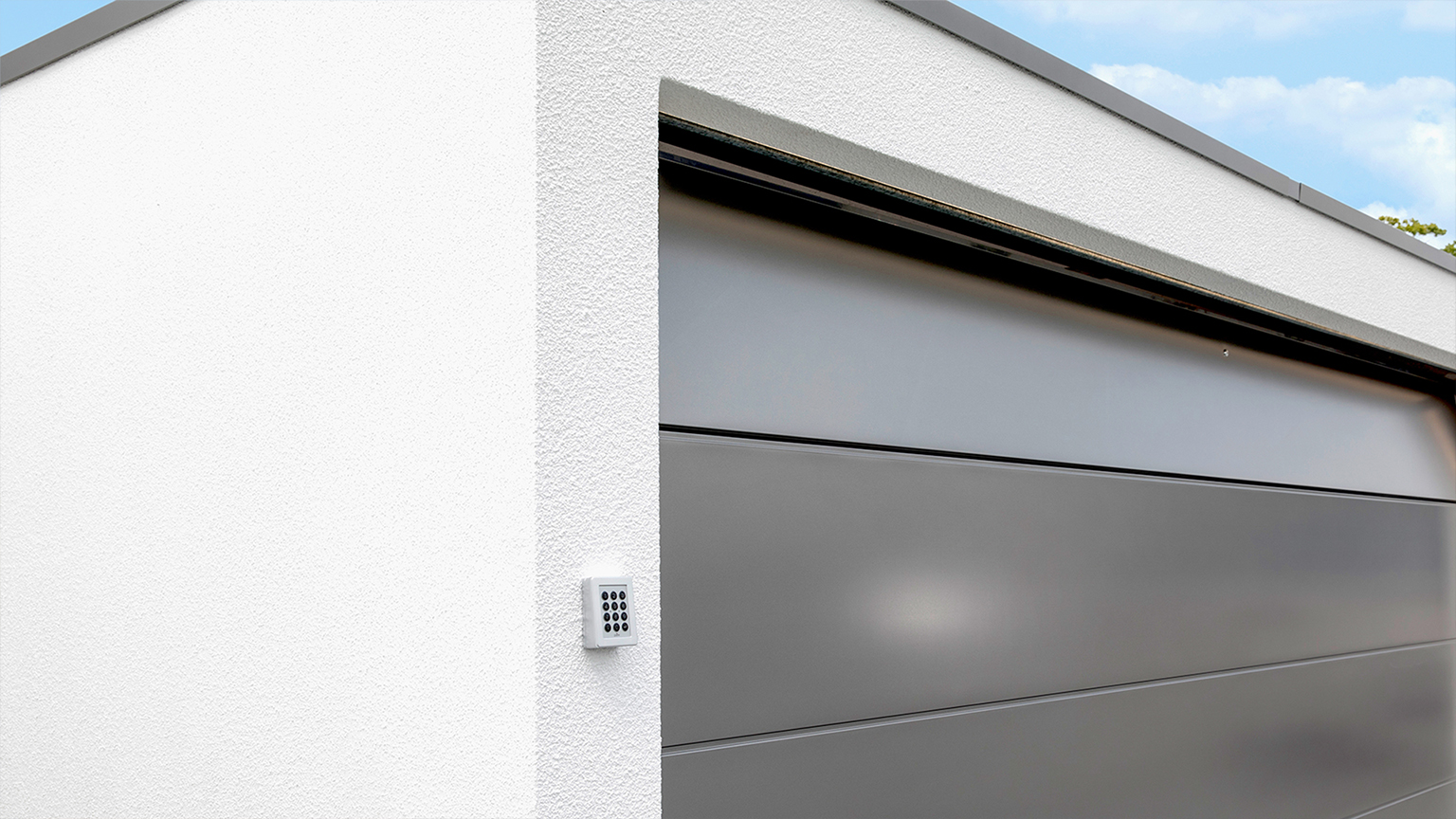 Porte sectionnelle avec clavier à code sur un garage préfabriqué moderne Vue rapprochée d’un garage préfabriqué blanc avec une porte sectionnelle grise et un clavier à code latéral pour une commande pratique de la porte par ZAPF