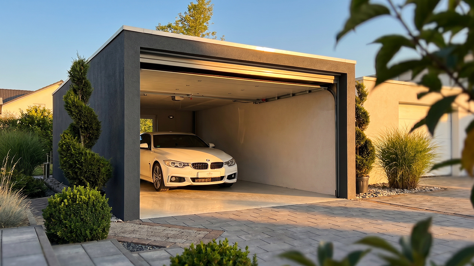 Garage préfabriqué en béton moderne avec porte sectionnelle ouverte Garage préfabriqué en béton gris avec toit plat, porte sectionnelle ouverte, voiture blanche à l’intérieur, allée pavée et aménagement paysager soigné de ZAPF Garages
