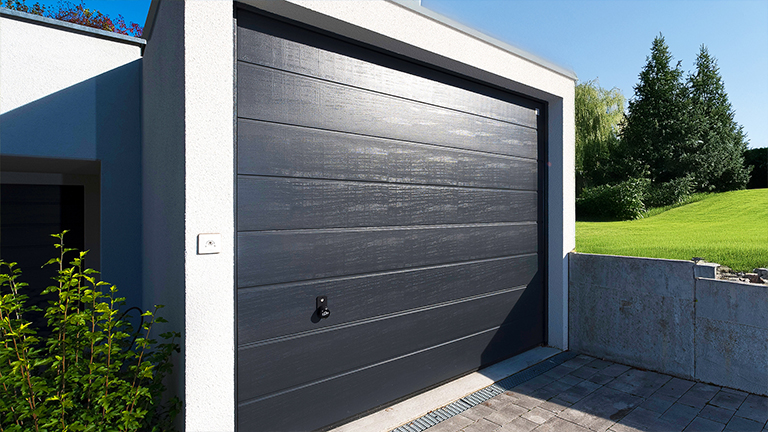 Moderne Betonfertiggarage mit hellem Baukörper, dunklem Sektionaltor in Holzoptik, gepflasterter Zufahrt und Grünfläche im Hintergrund von ZAPF